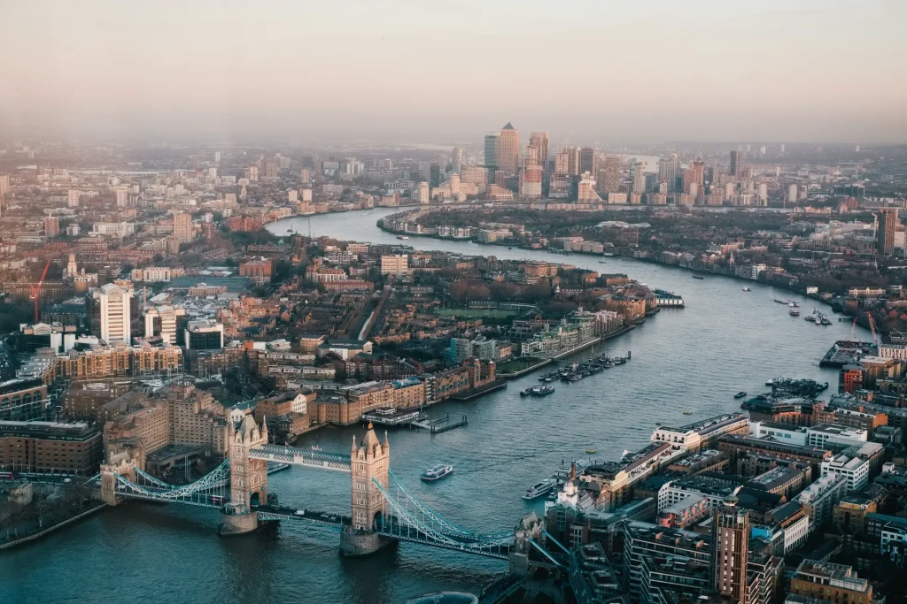 A sweeping aerial view of London during golden hour, showcasing the winding River Thames, the iconic Tower Bridge in the foreground, and the modern city skyline fading into the hazy distance.