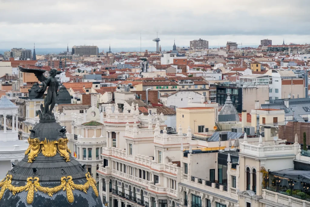 An expansive, elevated view over the historic rooftops of Madrid, featuring the ornate dark dome and golden-accented winged statue of the famous Metropolis Building prominently in the foreground.