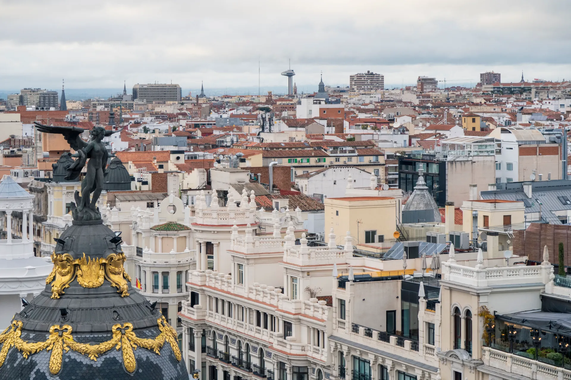 An expansive, elevated view over the historic rooftops of Madrid, featuring the ornate dark dome and golden-accented winged statue of the famous Metropolis Building prominently in the foreground.