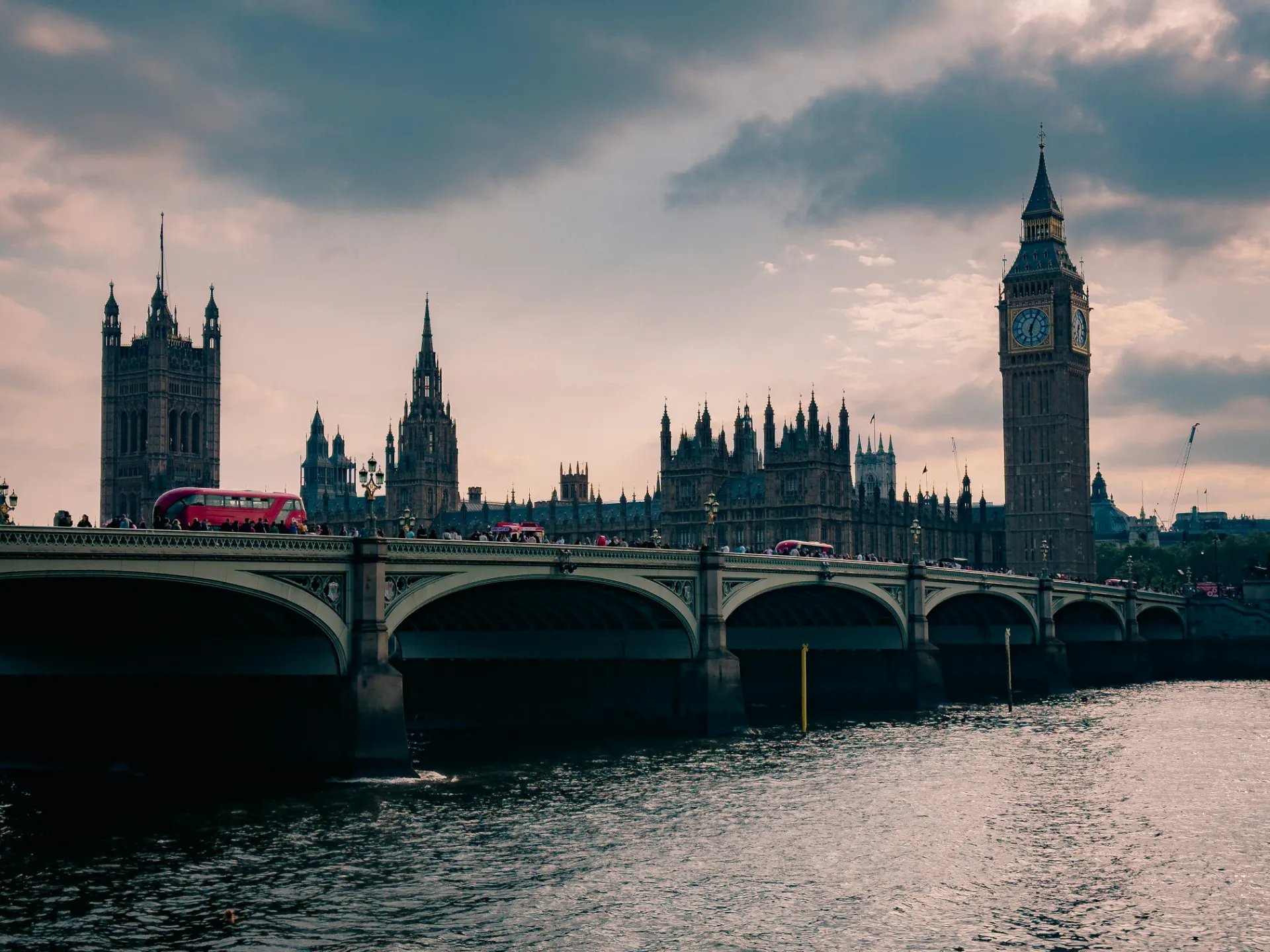 A moody, dramatic view of Big Ben and the Palace of Westminster under a cloudy, overcast sky, featuring a classic red double-decker bus crossing the arched Westminster Bridge.