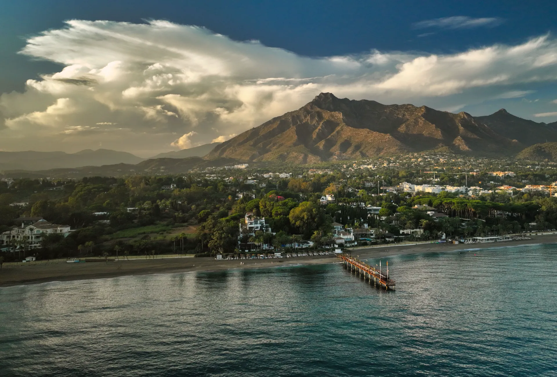 A scenic coastal view of the Costa del Sol at sunset, featuring a calm beach, a wooden pier stretching into the Mediterranean Sea, and lush green resorts backed by the majestic peak of La Concha mountain under a dramatic sky.