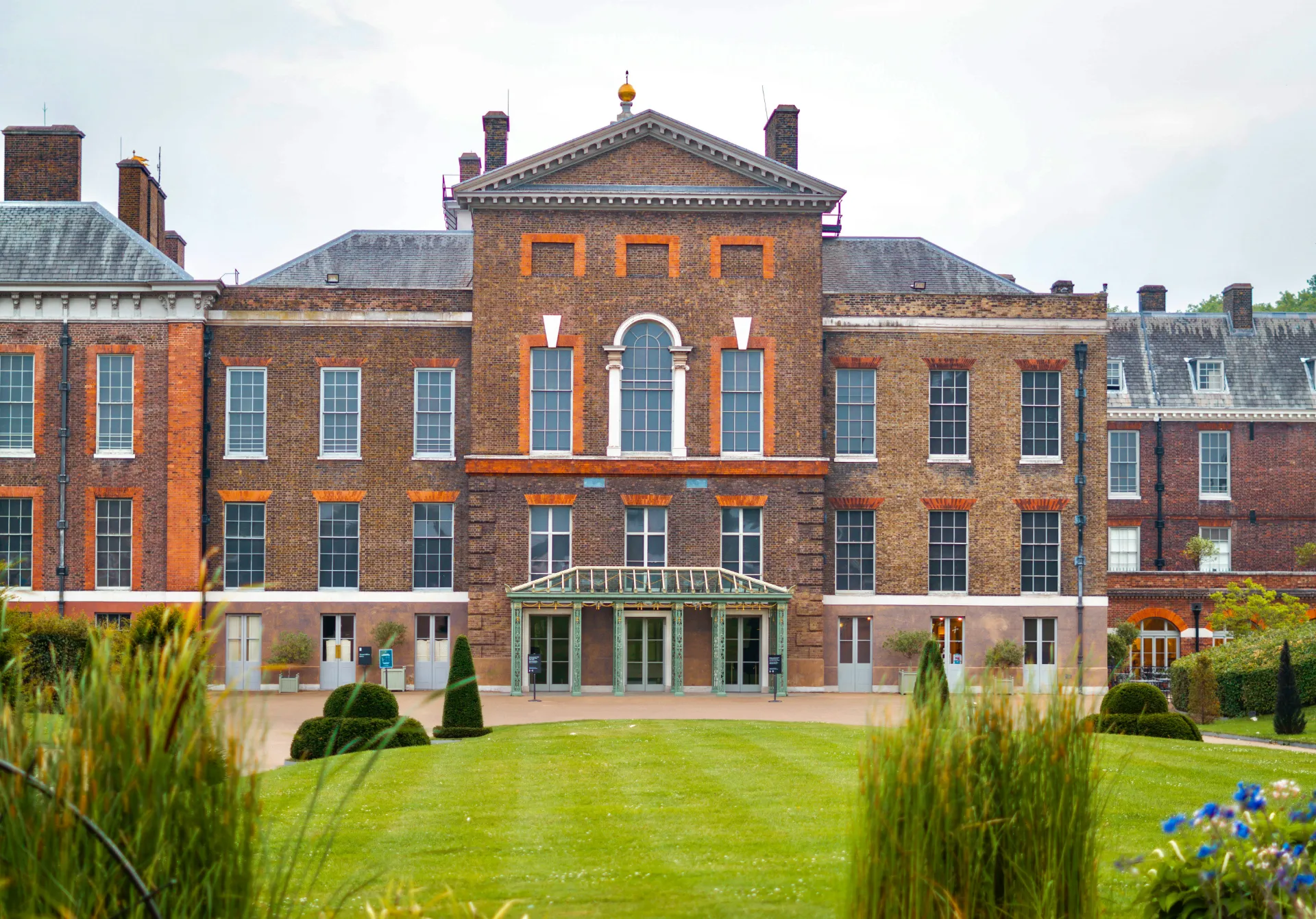 The grand brick exterior of Kensington Palace in London, featuring symmetrical rows of tall windows, a green entrance awning, and a neatly manicured green lawn in the foreground.