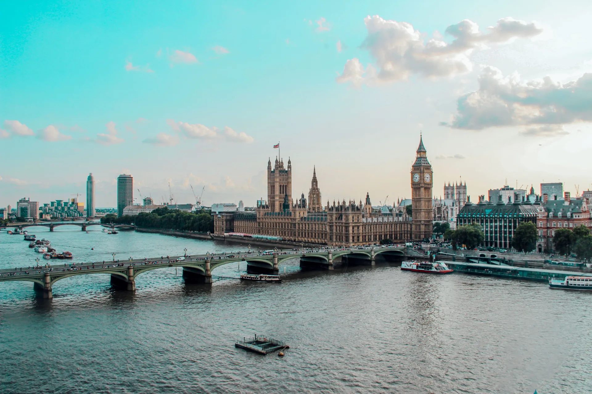 A wide scenic view of the Palace of Westminster and the iconic Big Ben clock tower sitting along the River Thames, with boats on the water and Westminster Bridge spanning the foreground under a bright blue sky.