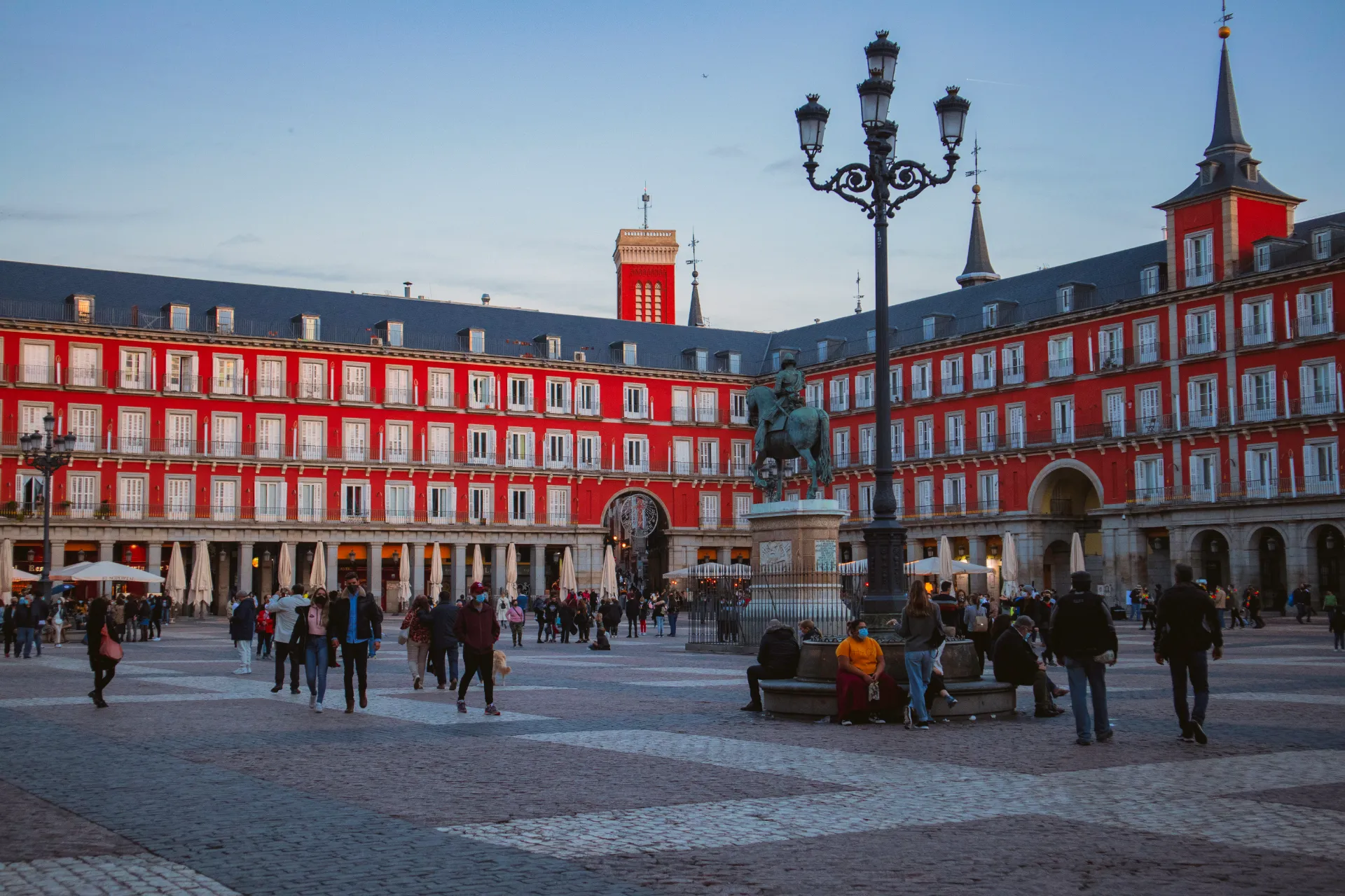 The historic Plaza Mayor square in Madrid bustling with pedestrians, characterized by striking red buildings with continuous ground-floor arcades, wrought-iron balconies, and a central bronze equestrian statue of King Philip III.