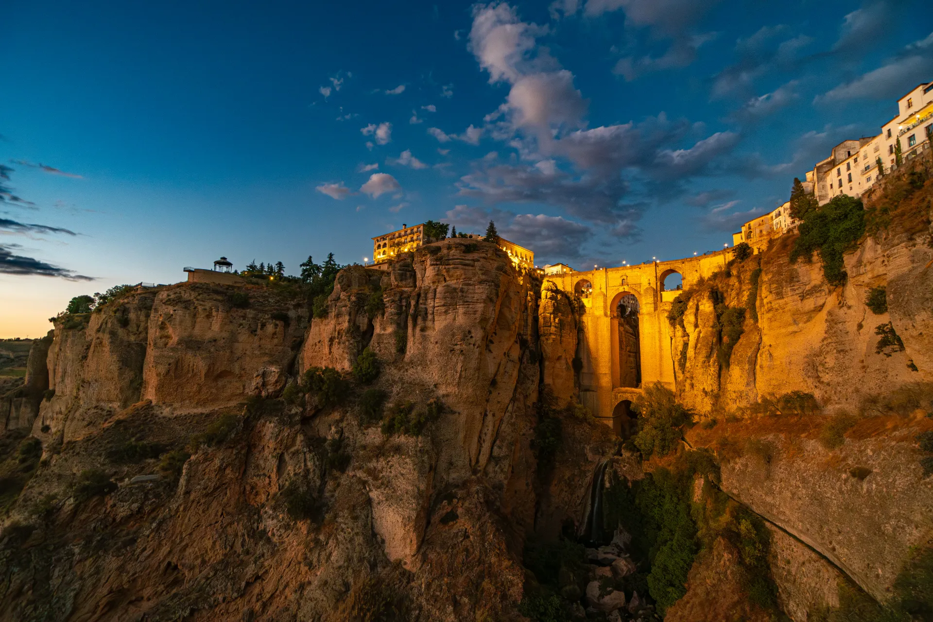 A breathtaking twilight view of the massive stone arches of the Puente Nuevo bridge in Ronda, Spain. The bridge spans a deep, dramatic rocky gorge, with traditional white buildings perched right on the edge of the illuminated cliffs.