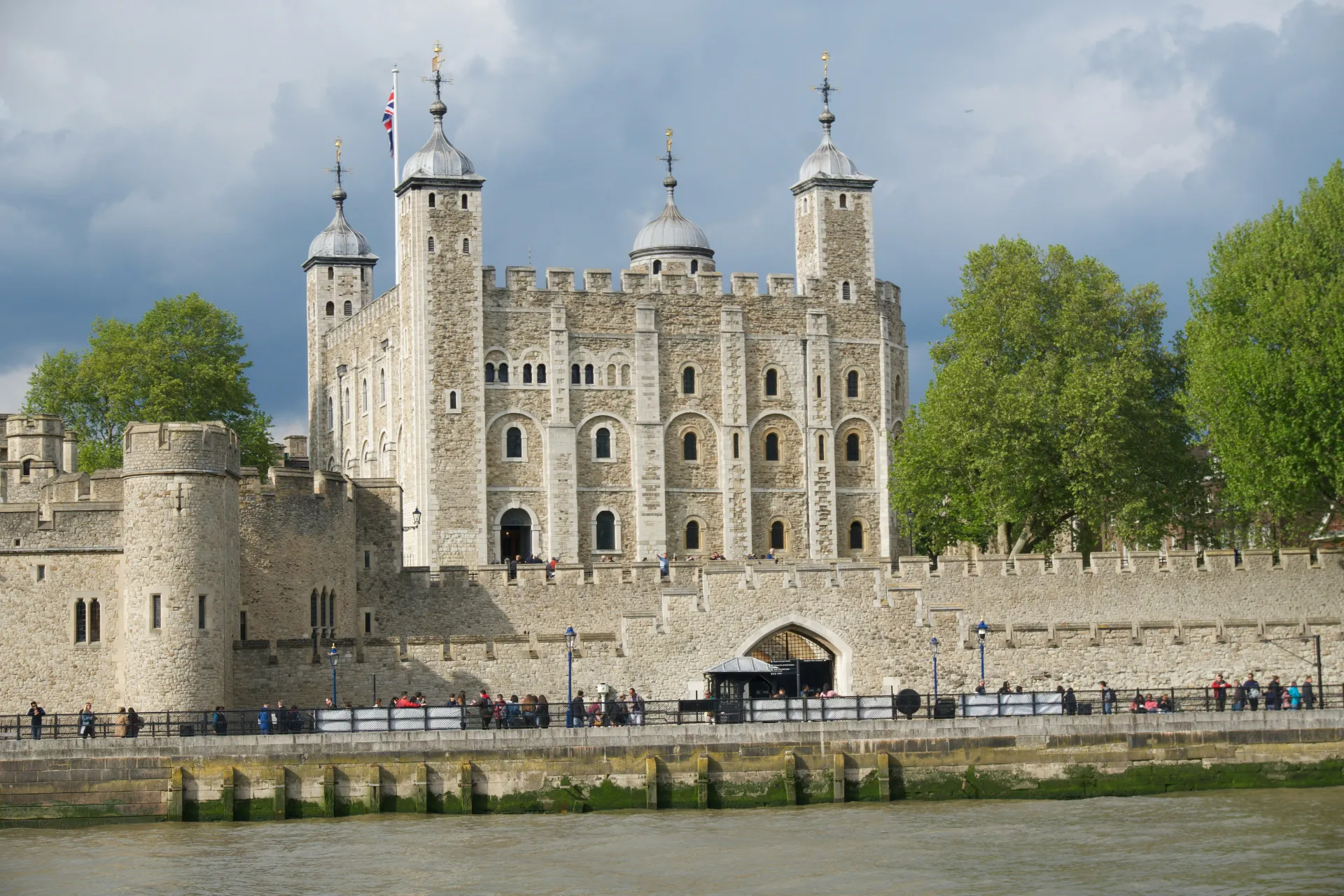 A daytime view of the historic Tower of London, featuring the central stone fortress with its four domed turrets and the Union Jack flying, surrounded by an outer stone wall where visitors are walking.