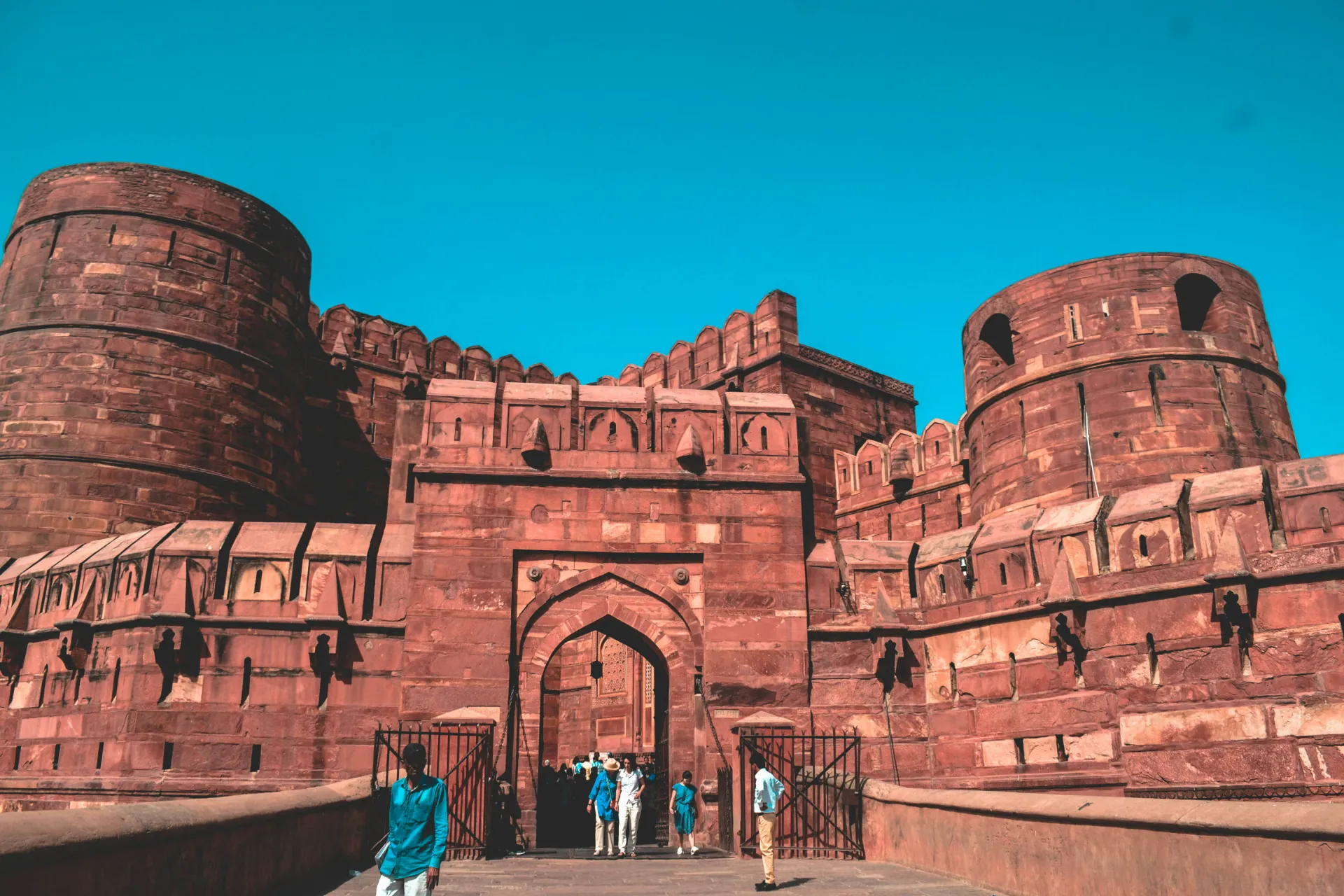 Imposing red sandstone walls and circular towers of Agra Fort with visitors walking across the entry bridge toward the grand arched gateway.