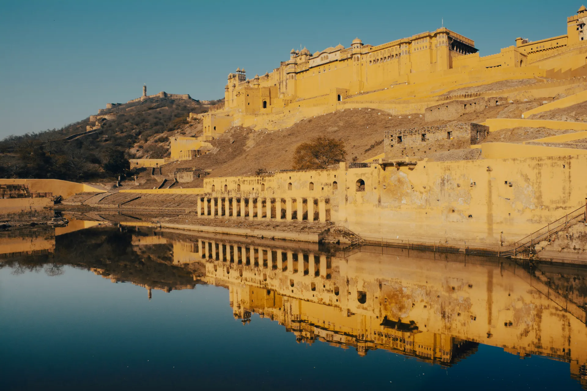 The expansive yellow sandstone Amer Fort in Jaipur perched on a hillside, reflecting in the calm water below under a clear sky.