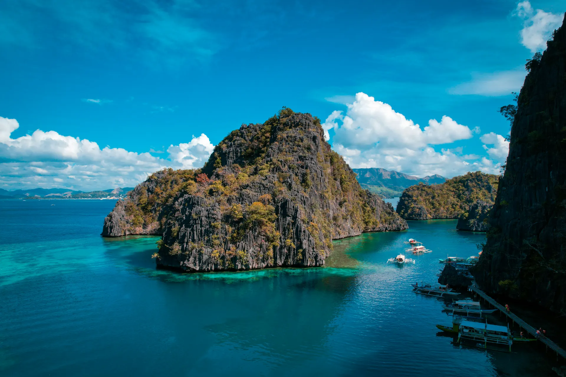 An elevated view of dramatic limestone karst formations covered in green foliage, rising above clear turquoise ocean waters with traditional outrigger boats anchored along the shore.