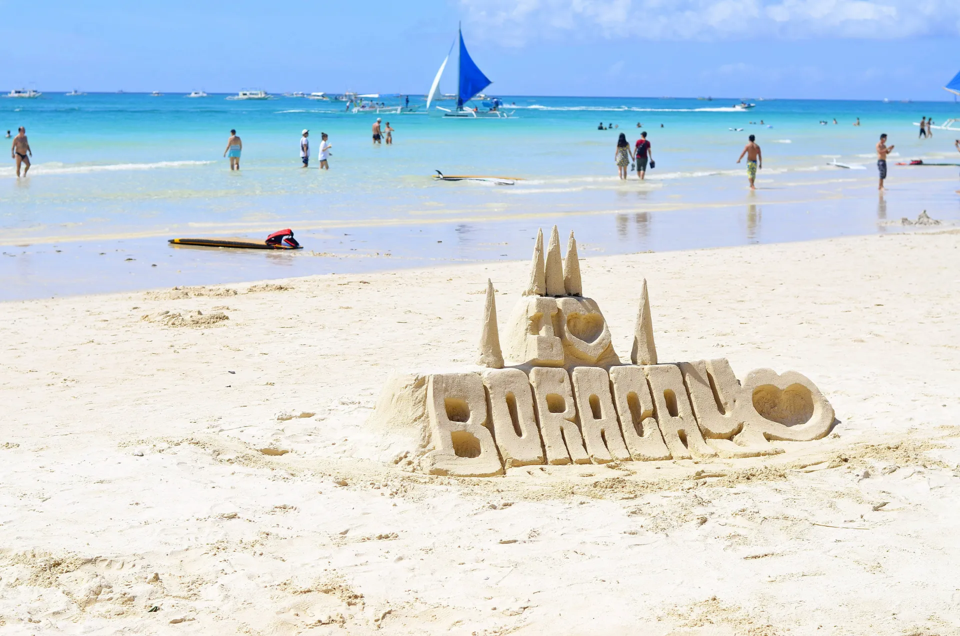 A sandcastle carved on a white sandy beach with text reading "I LOVE BORACAY," set against clear turquoise water, sailboats, and beachgoers under a sunny sky.
