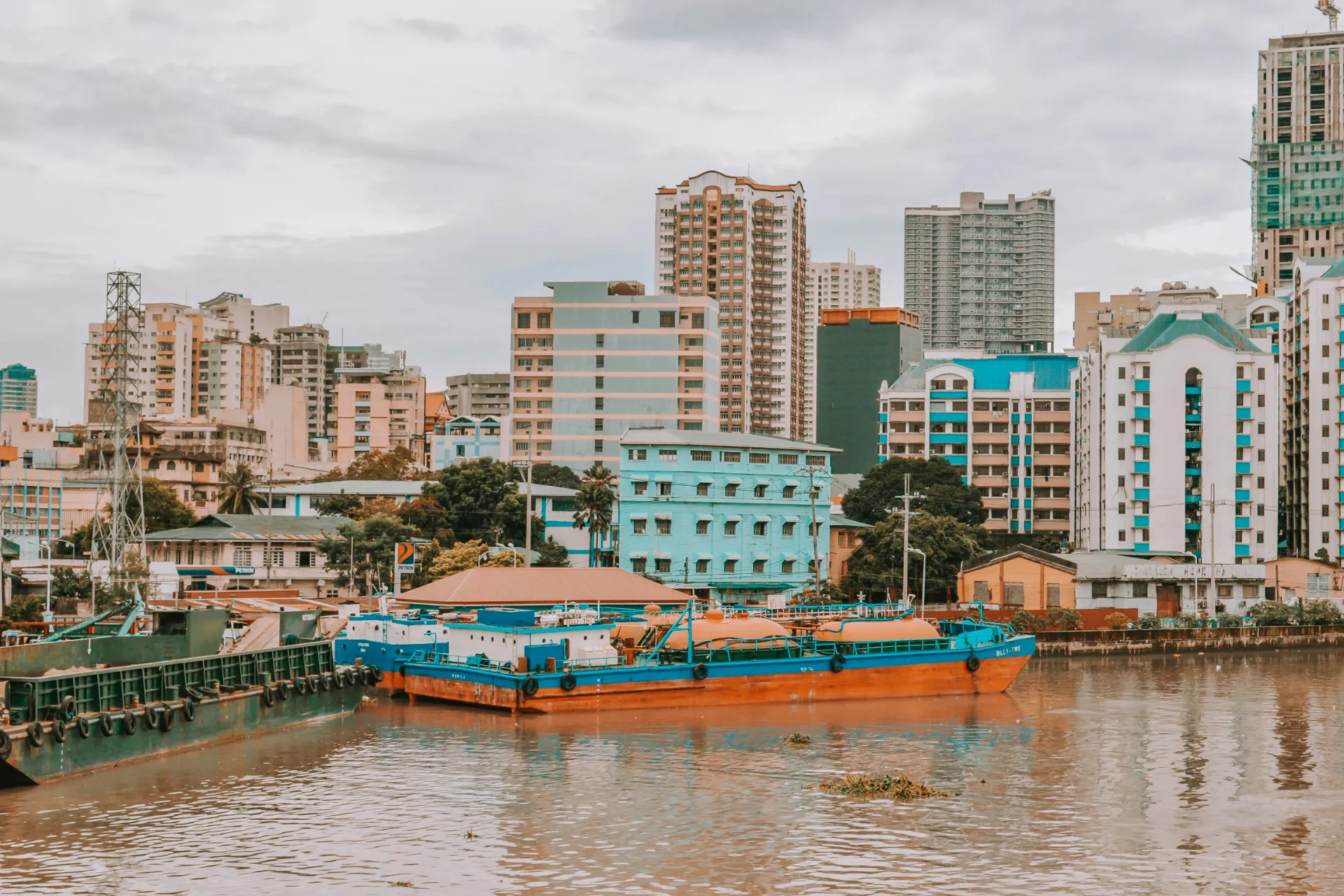 A view across the Pasig River in Manila showing blue and orange cargo barges moored in the water, with a dense skyline of colorful residential and commercial buildings under a cloudy sky.