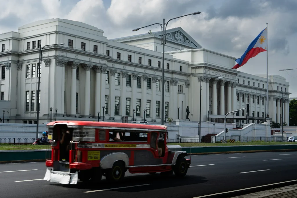 A classic red and silver Philippine Jeepney driving along an urban street with the grand, white Neoclassical National Museum building and the Philippine flag flying in the background.