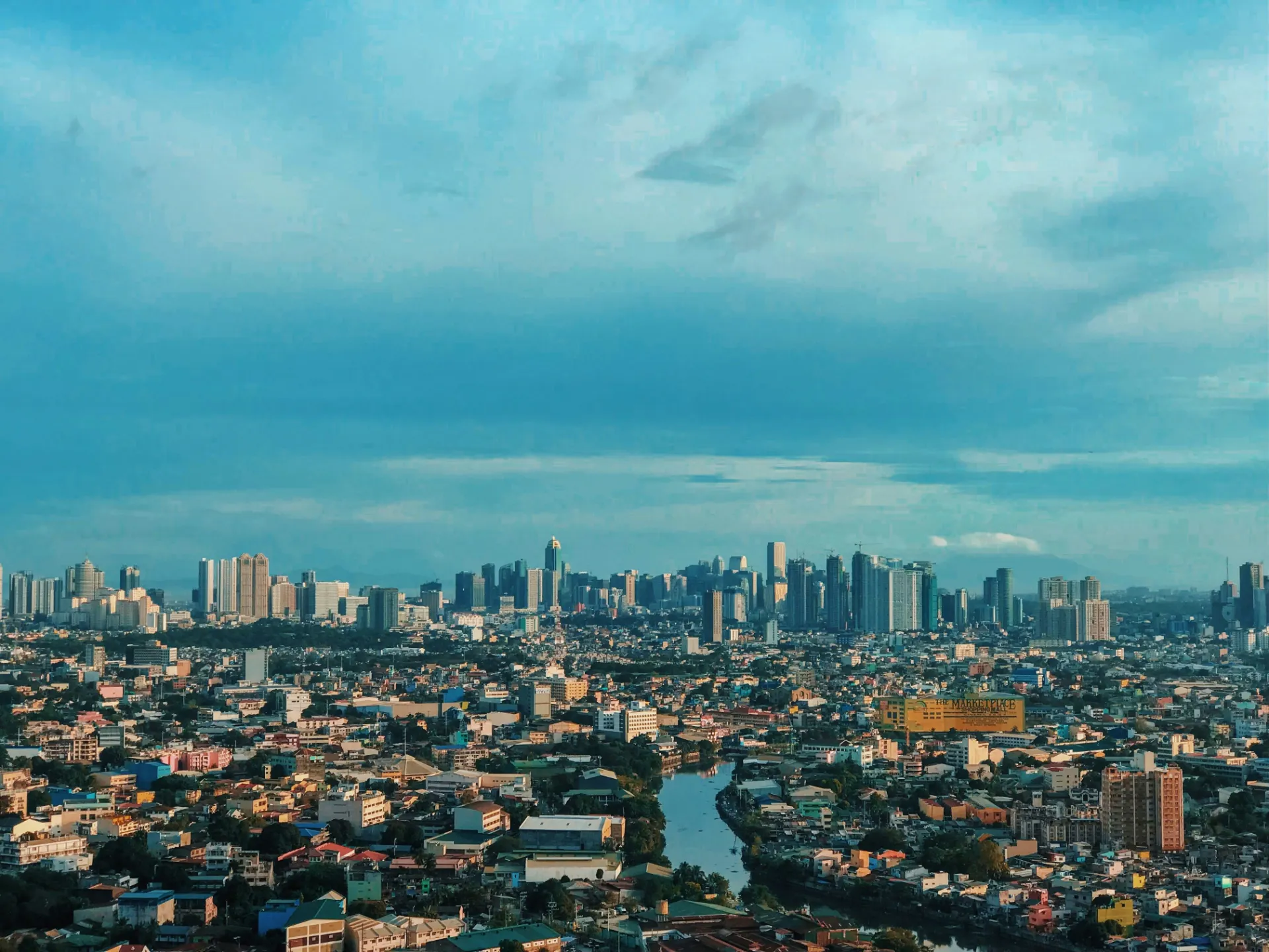 A sweeping aerial view over Metro Manila showing dense neighborhood rooftops, a winding river, and a modern skyscraper skyline beneath a wide blue sky.