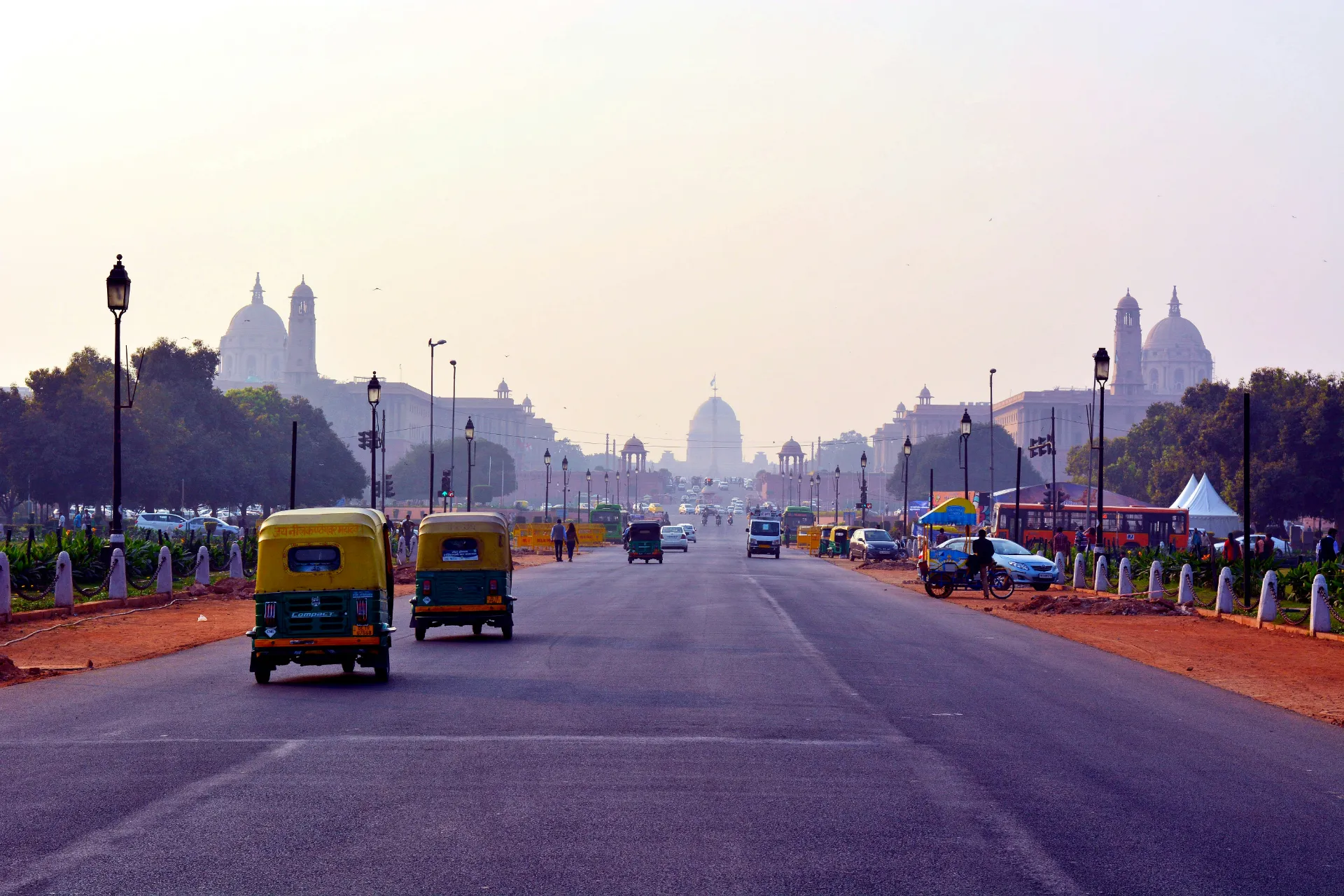 A wide view looking down Kartavya Path in New Delhi with yellow auto-rickshaws driving toward the grand Secretariat buildings in the hazy distance.